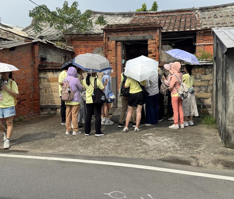學員圍聚在紅磚古厝門前，聆聽這座百年建築的故事。斑駁的燕尾磚牆與瓦頂，訴說著八里聚落曾經的生活痕跡。（照片提供者：新北市教育局 / 拍攝日期：2026.08.11）